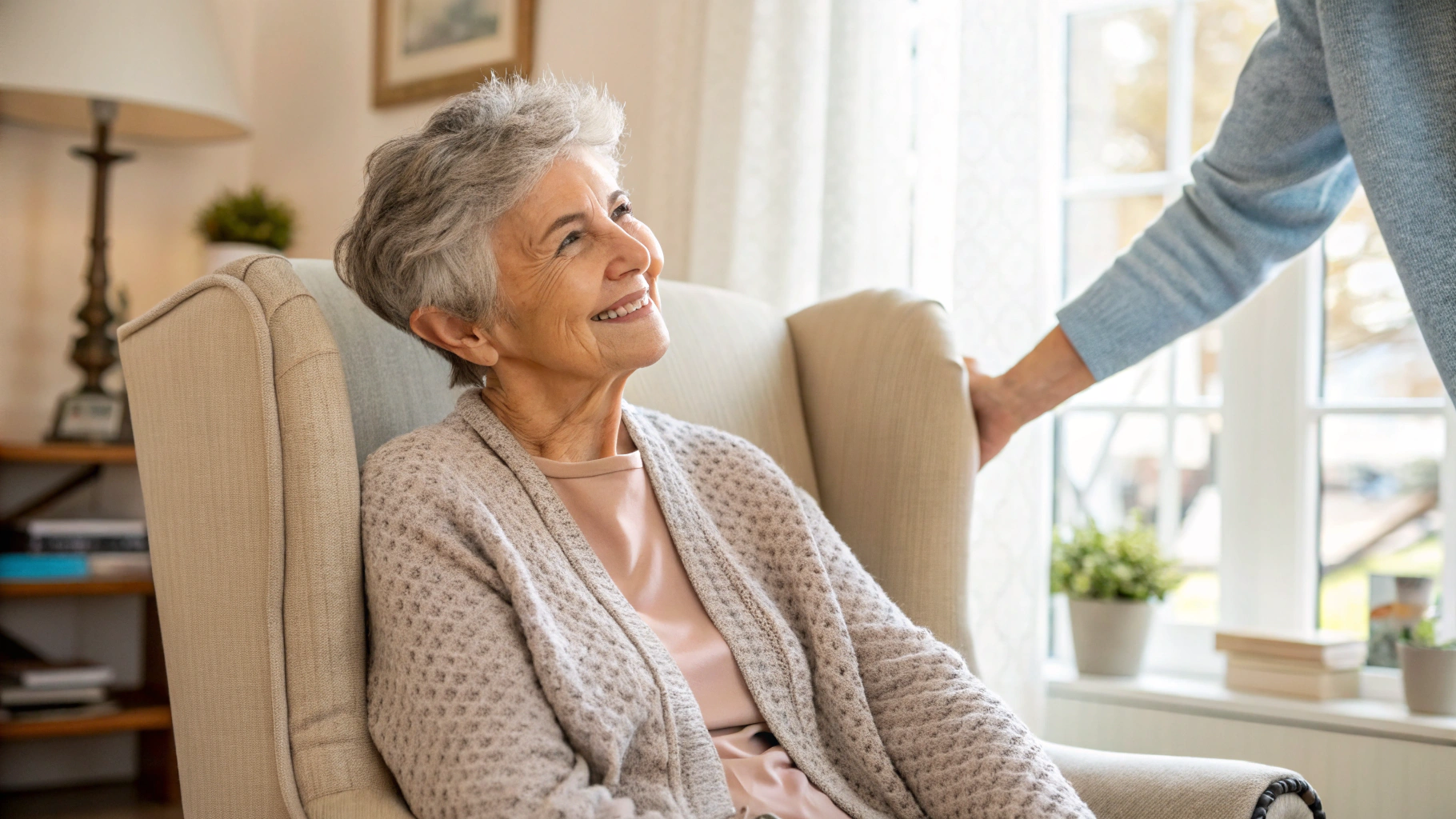 A smiling senior woman in an armchair at home looking up at a person standing beside her